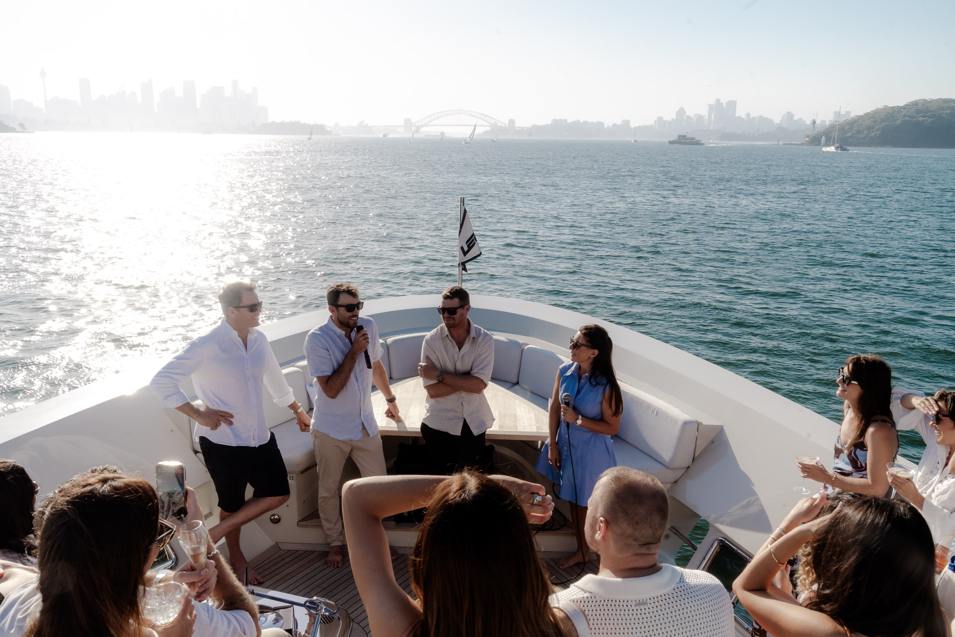 A man speaks into a microphone to seated guests on a superyacht deck, with the Sydney skyline and Harbour Bridge glowing behind.