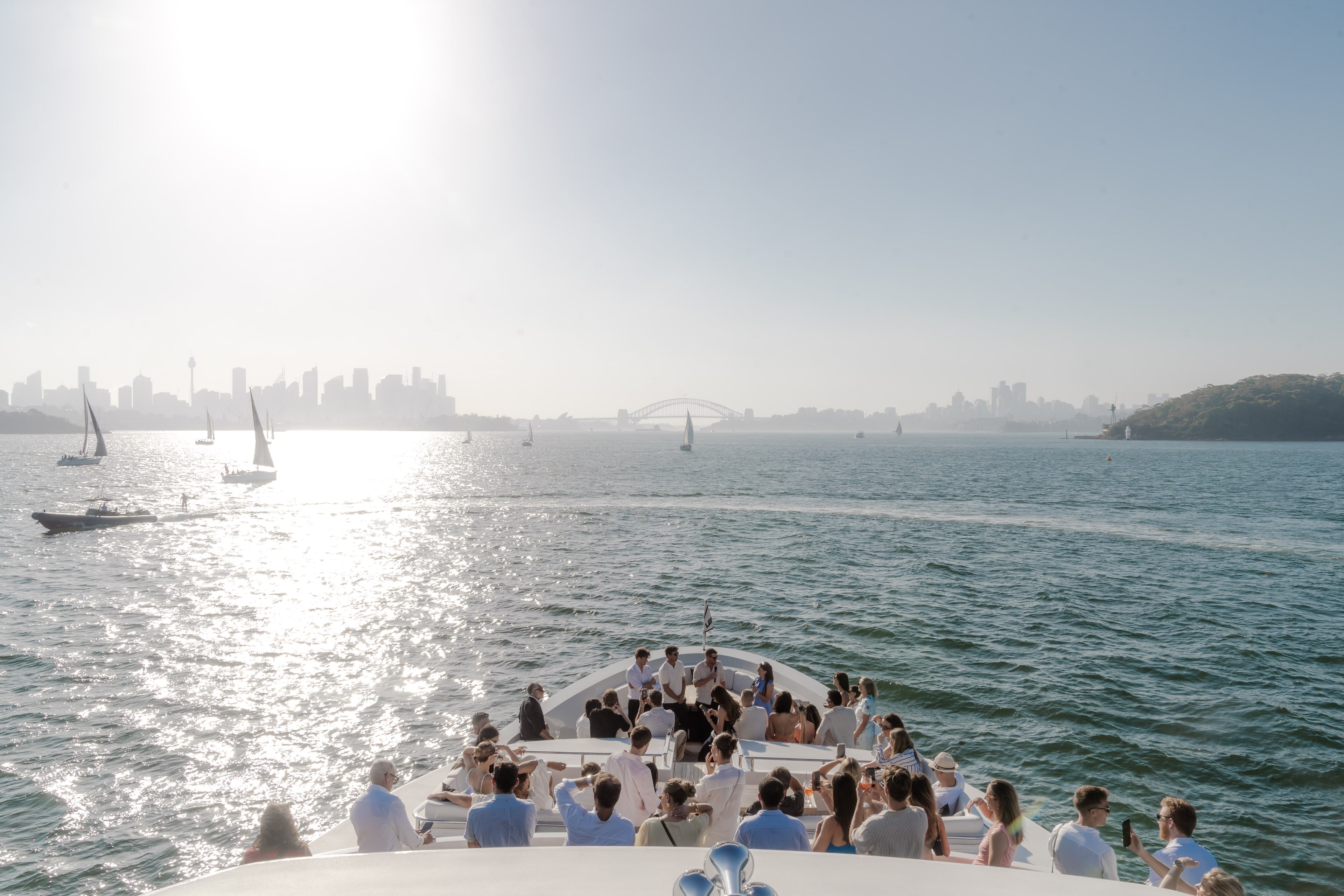 Guests in white gather on a superyacht deck watching sailboats on Sydney Harbour, with the city skyline and Harbour Bridge backlit by sun.