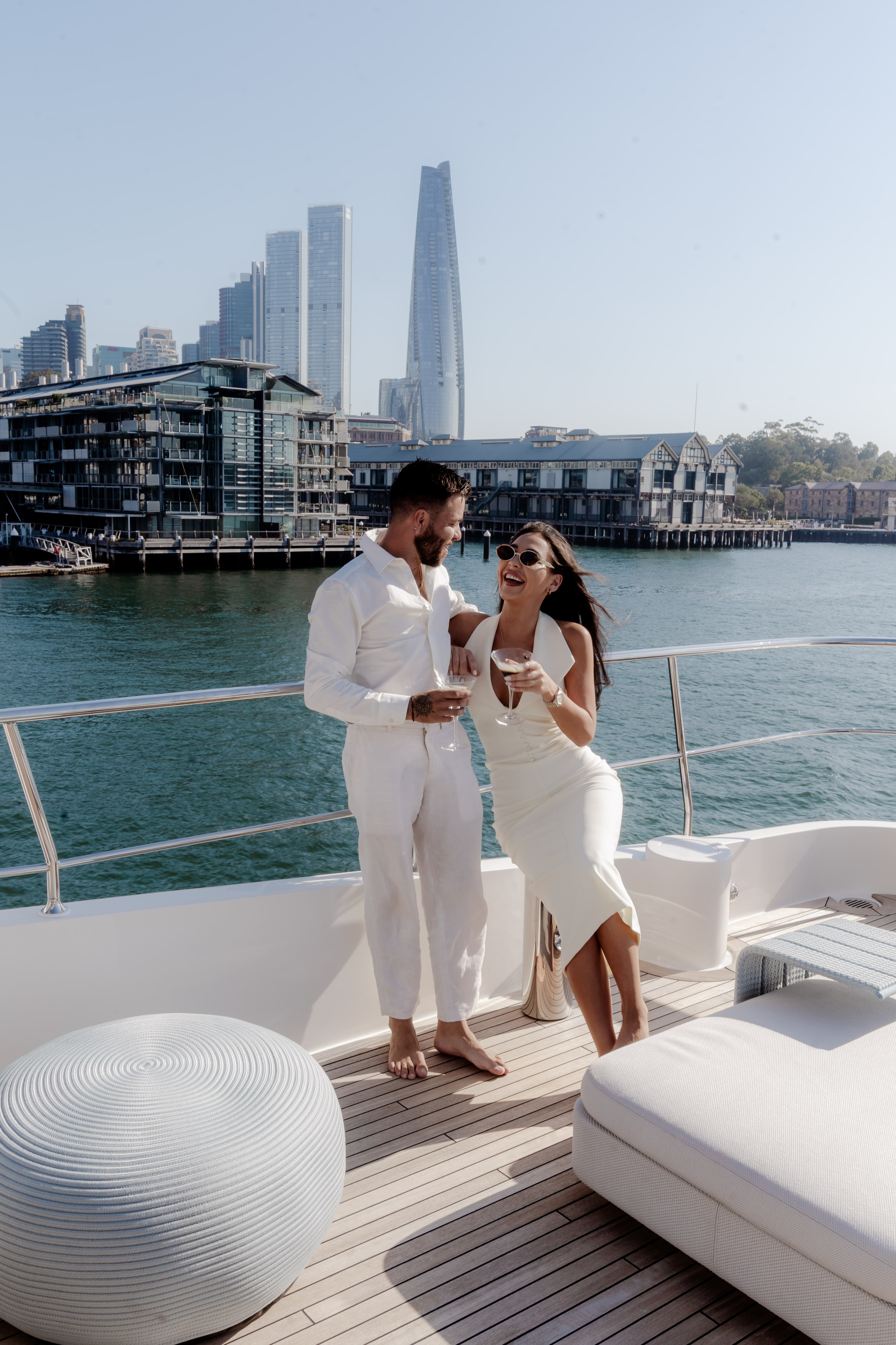 A couple dressed in white laugh together with drinks on a superyacht deck, with the Sydney city skyline and Barangaroo behind them.