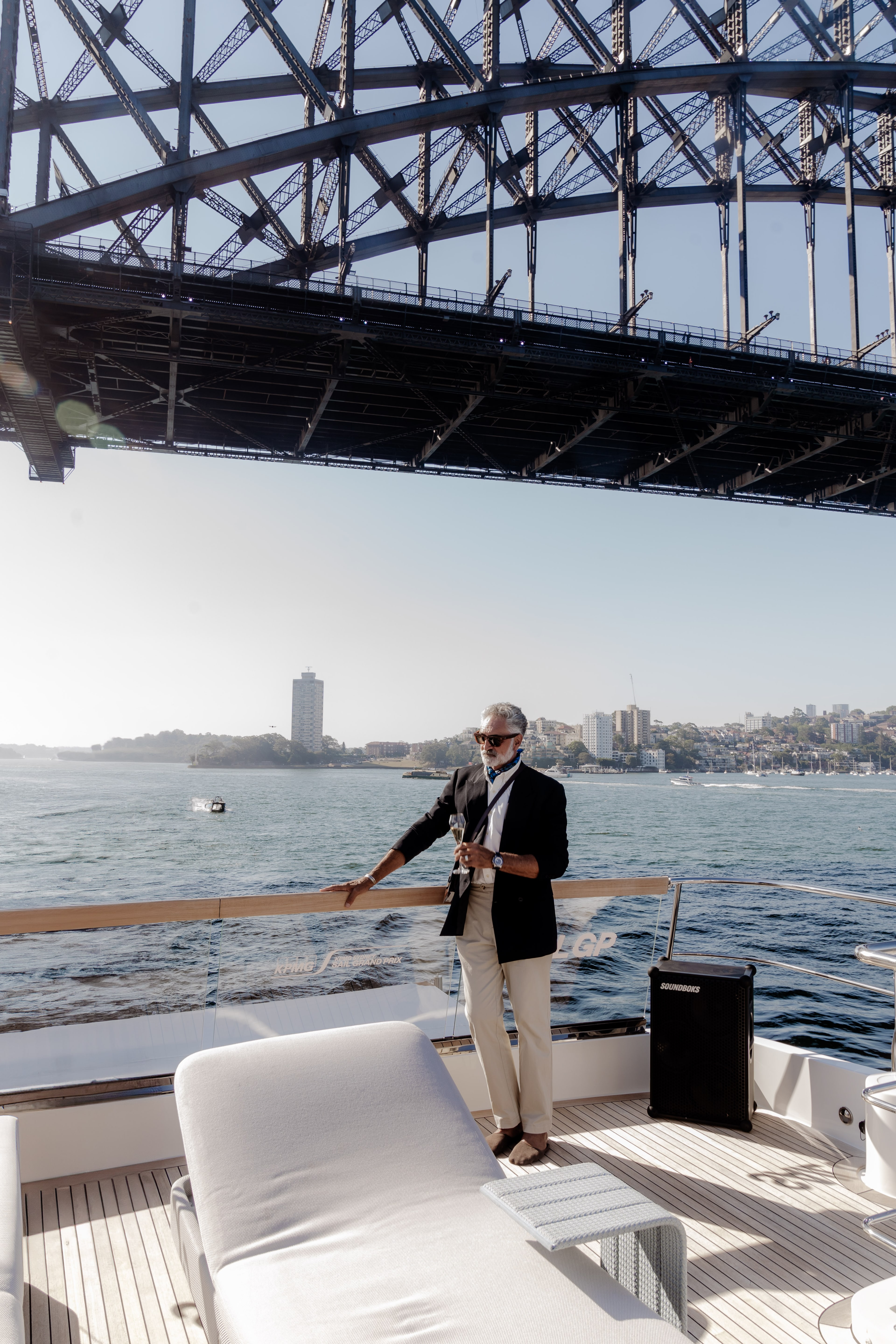 A saxophonist in a dark blazer performs on a superyacht deck directly beneath the Sydney Harbour Bridge on a sunny day.