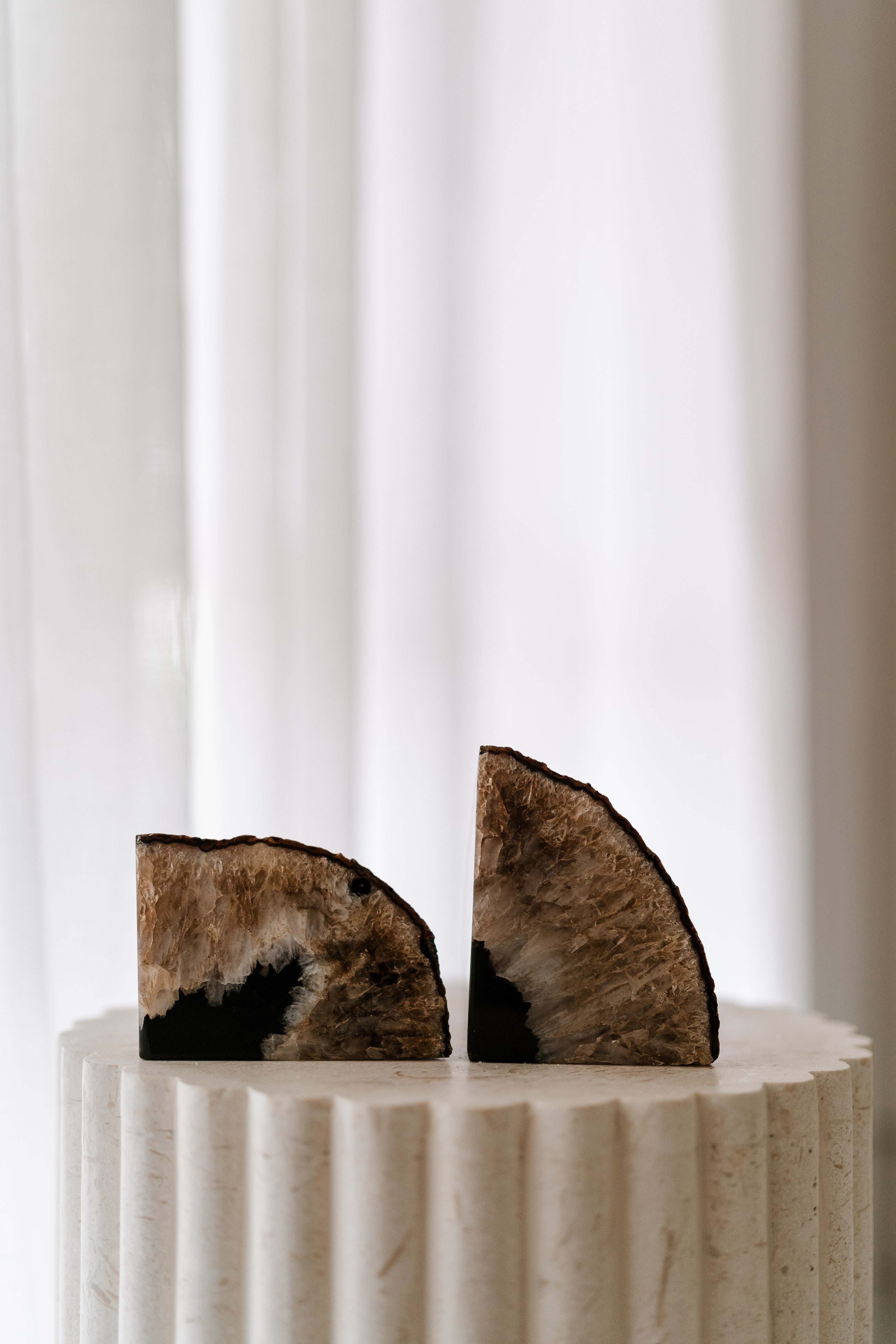 Pair of brown agate crystal bookends with raw crystalline centres displayed on a fluted stone pedestal by a bright curtain.