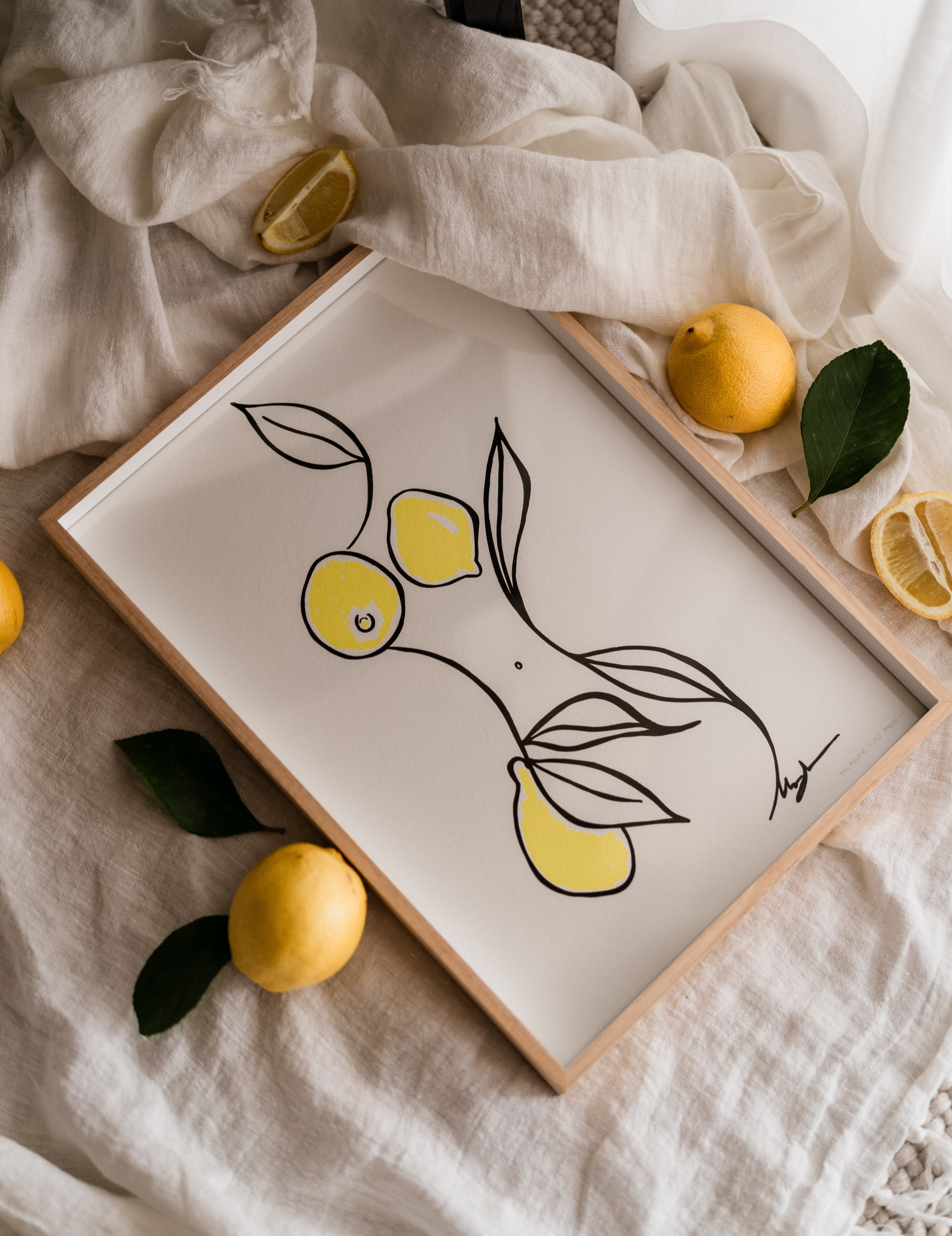 Framed minimalist black-line lemon illustration styled on crumpled linen with fresh and halved lemons and green leaves.
