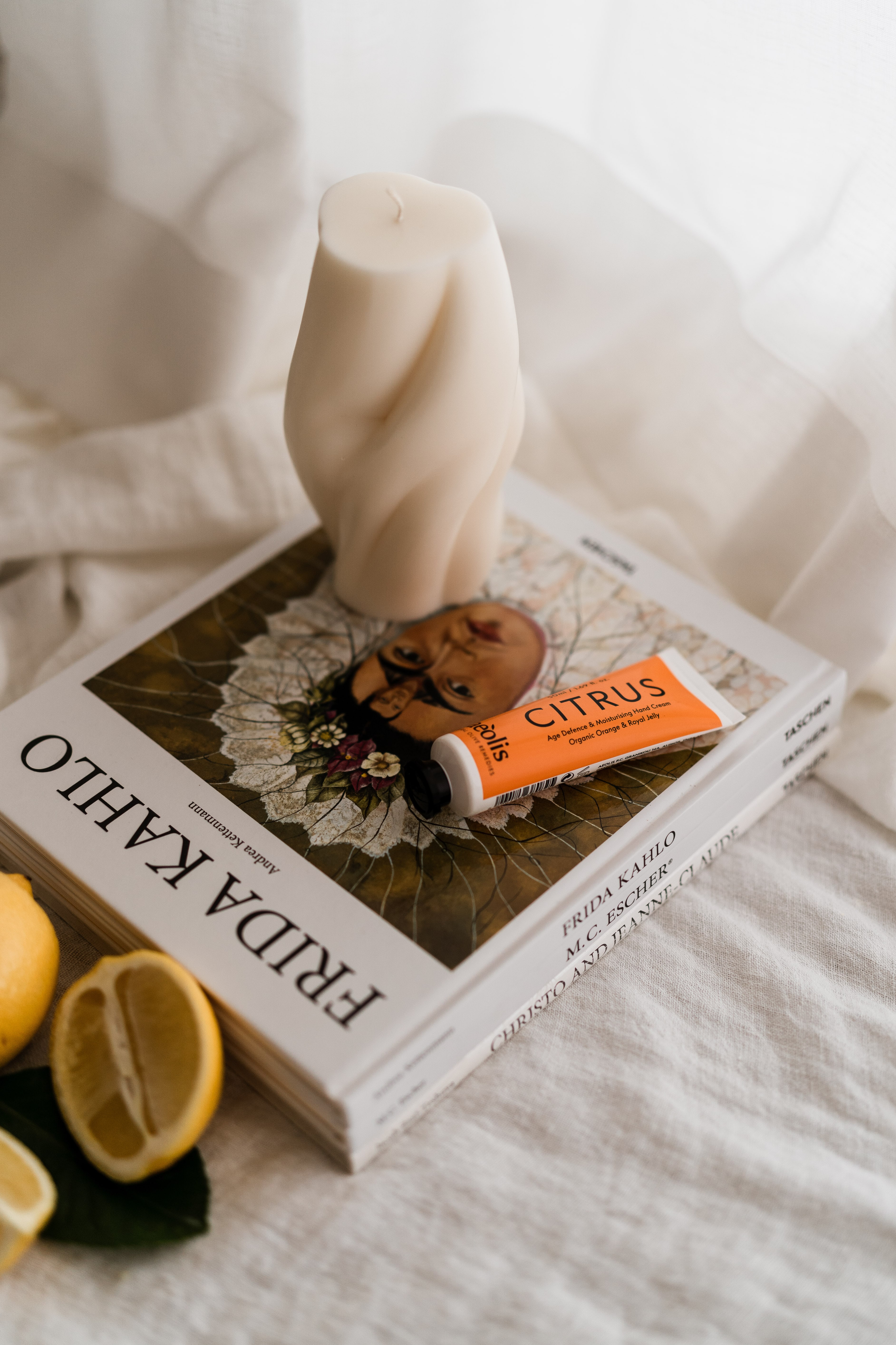Cream twisted candle on a stack of Frida Kahlo and Taschen art books with an orange Citrus hand cream tube and lemons on linen.