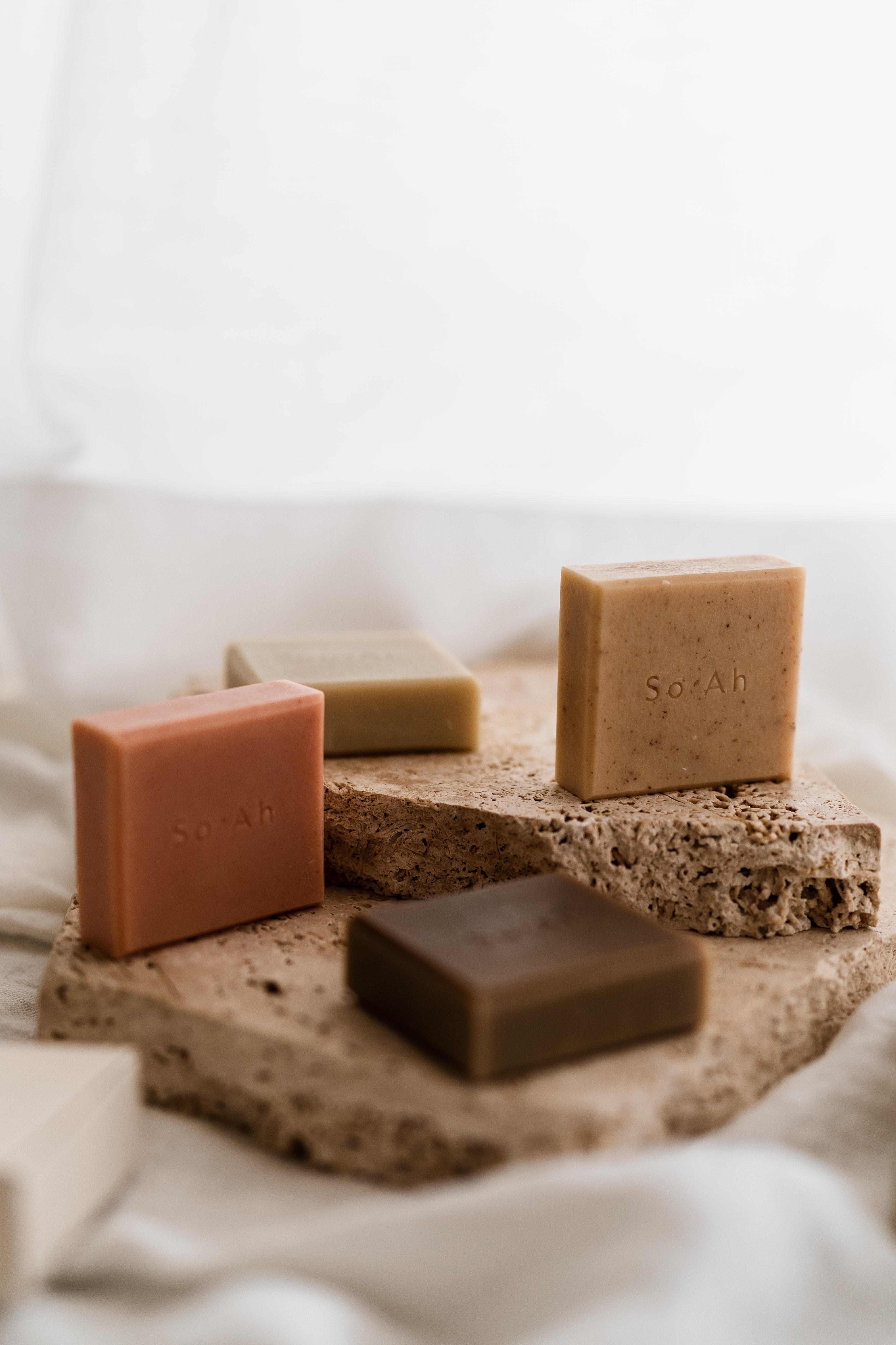 Four So-Ah embossed artisan soap bars in terracotta, olive and brown tones arranged on a raw travertine slab over linen.