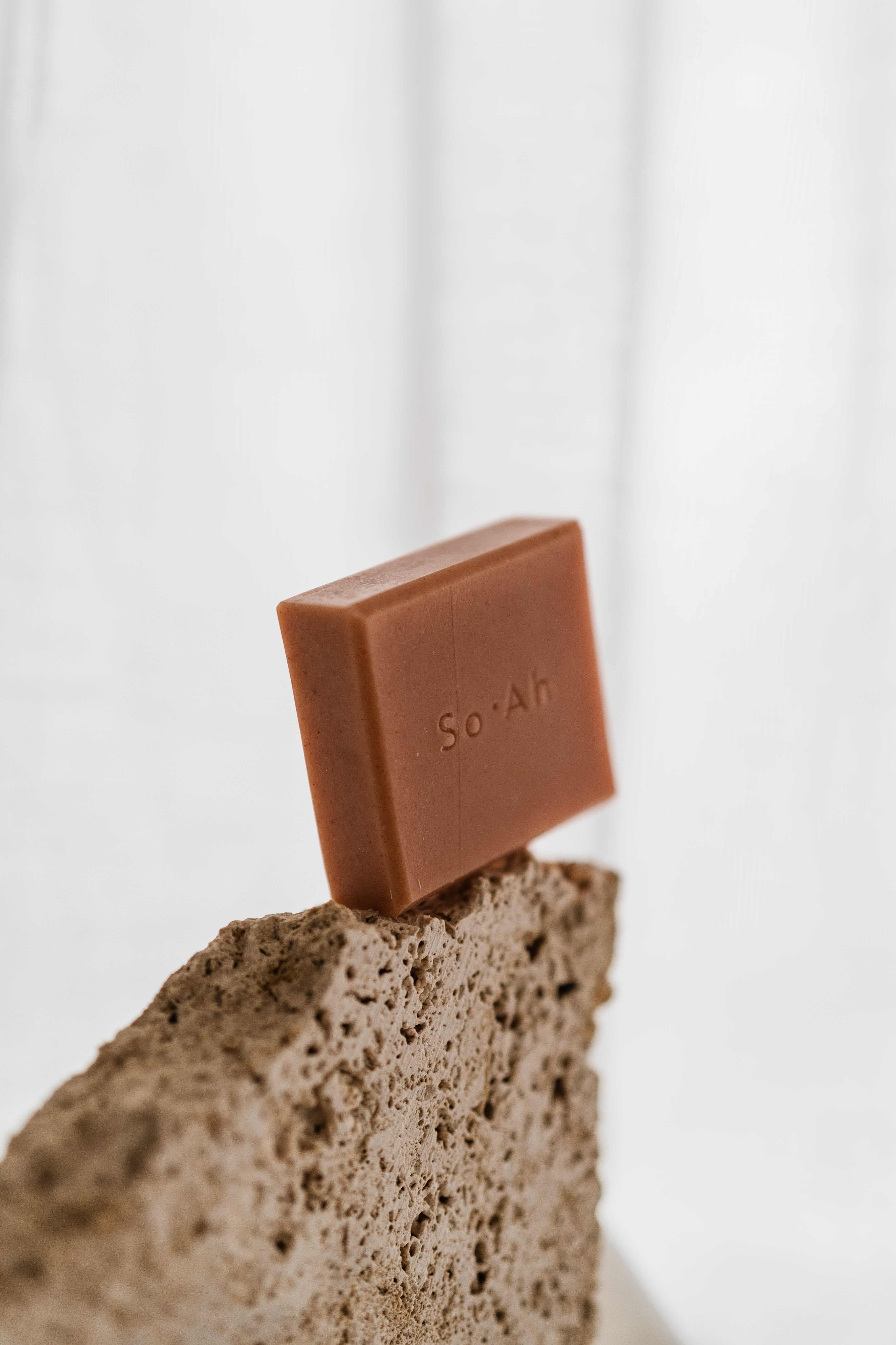 Single terracotta So-Ah soap bar balanced on the edge of a porous travertine stone against a soft white backdrop.