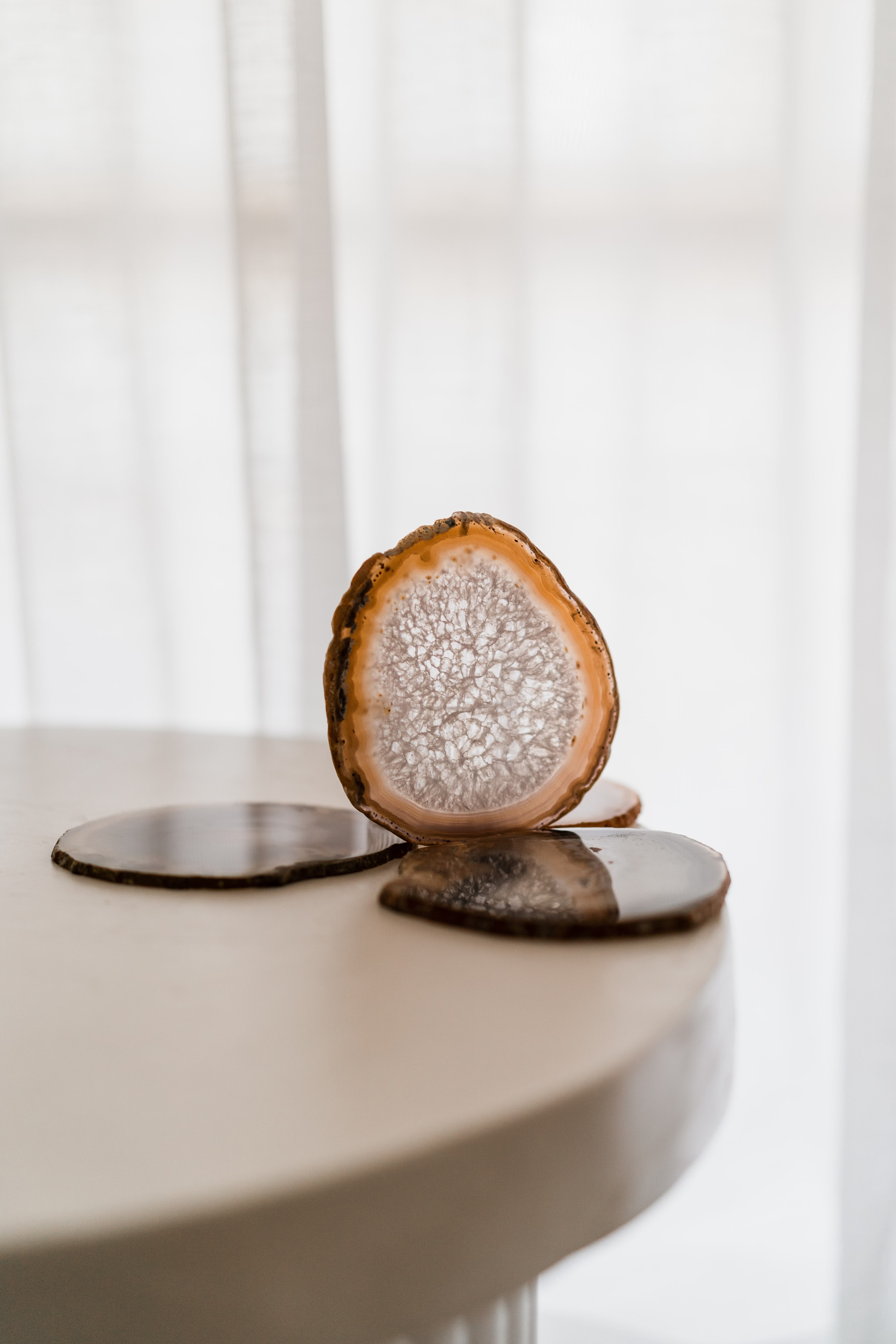 Sliced agate coasters with crystalline centres, one standing upright, arranged on a cream round table by sheer curtains.