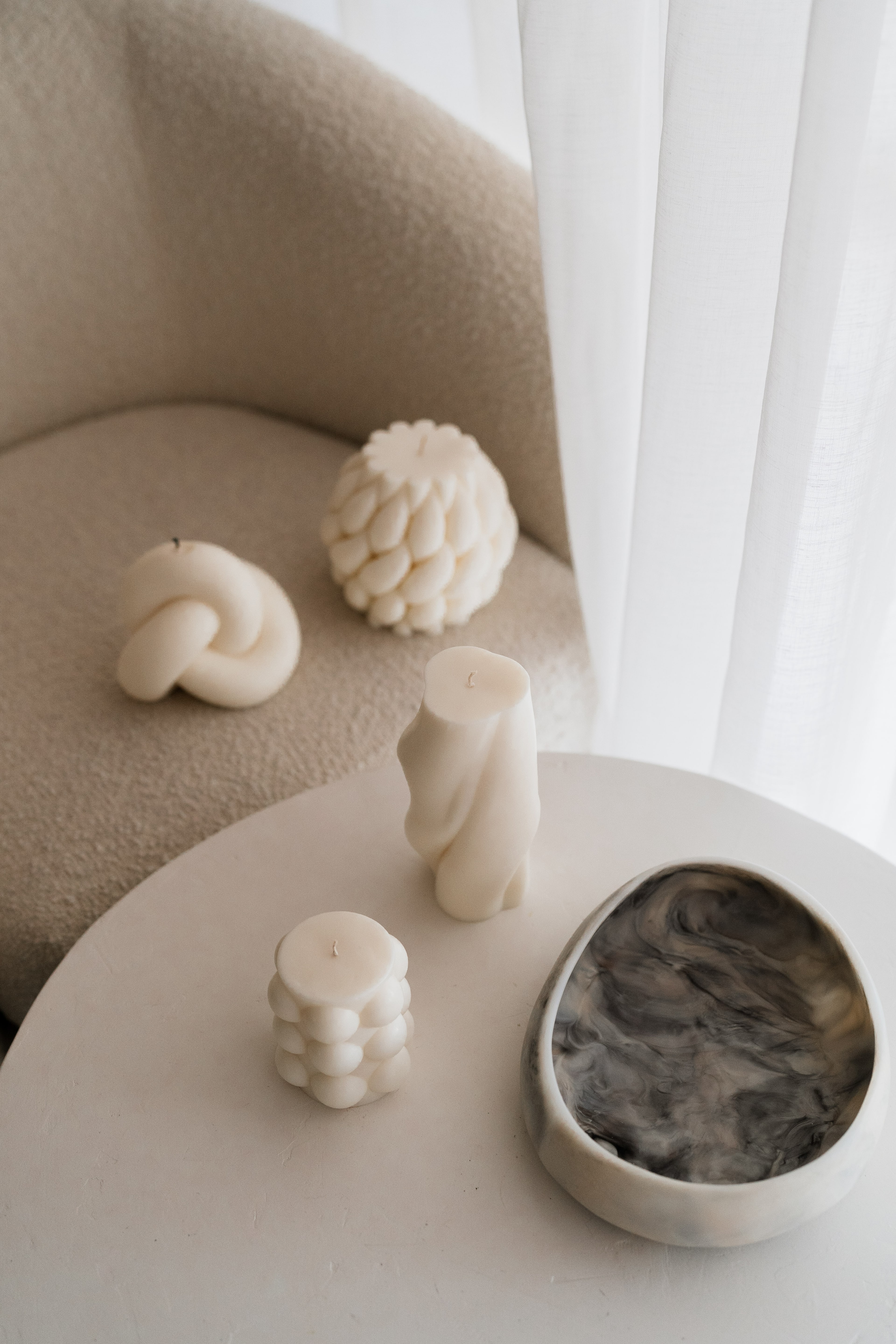 Sculptural cream candles including knot, pinecone and bobble shapes with a black marbled trinket dish on a boucle chair and side table.