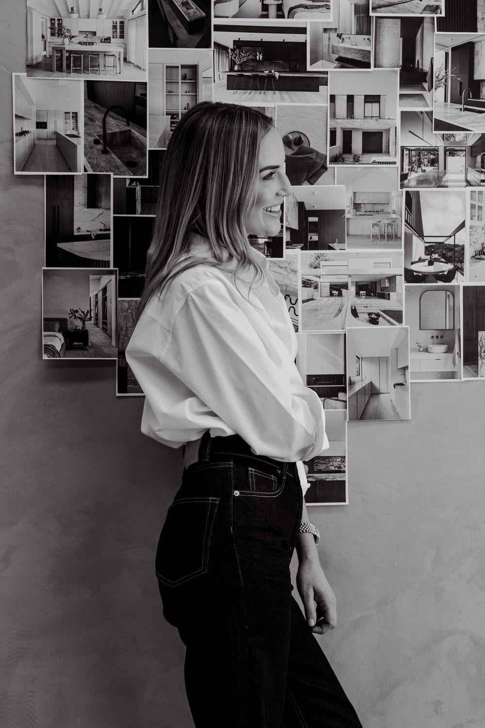 Black-and-white profile of a smiling woman in a white shirt and dark jeans standing before a wall covered in interior design photographs.