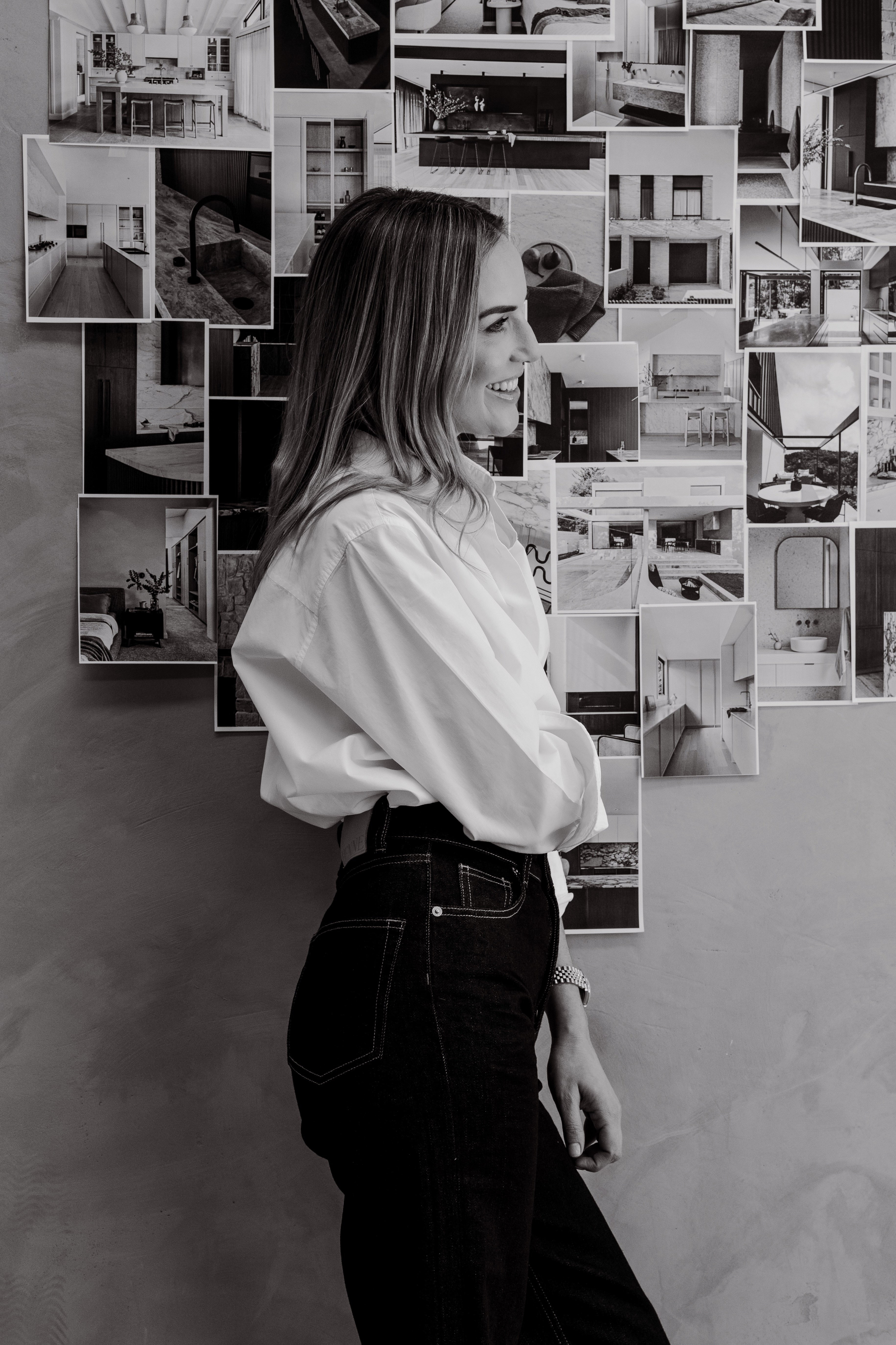 Black-and-white profile of a smiling woman in a white shirt and dark jeans standing before a wall covered in interior design photographs.