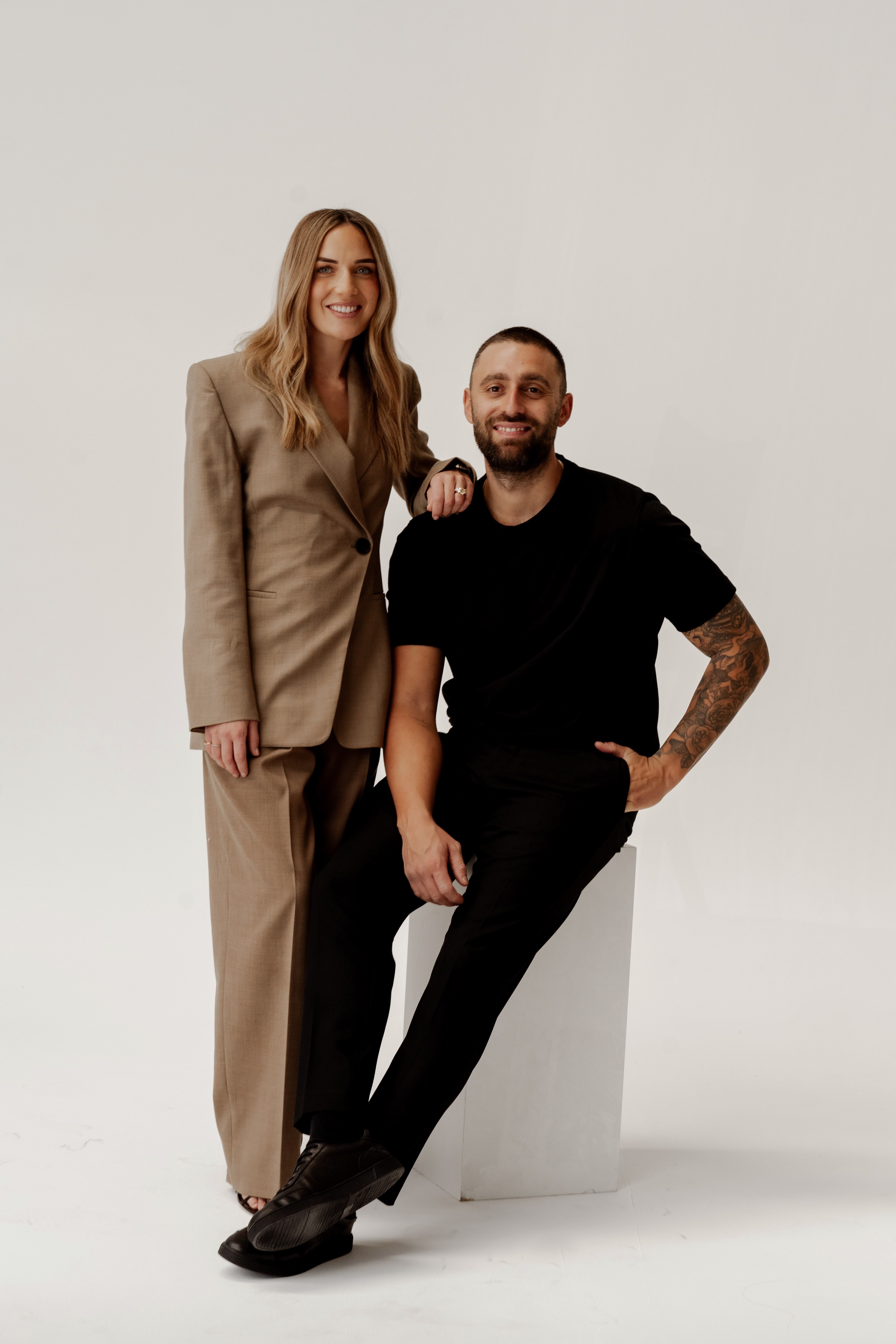 Studio portrait of a smiling woman in a beige suit standing beside a tattooed man in black seated on a white plinth.