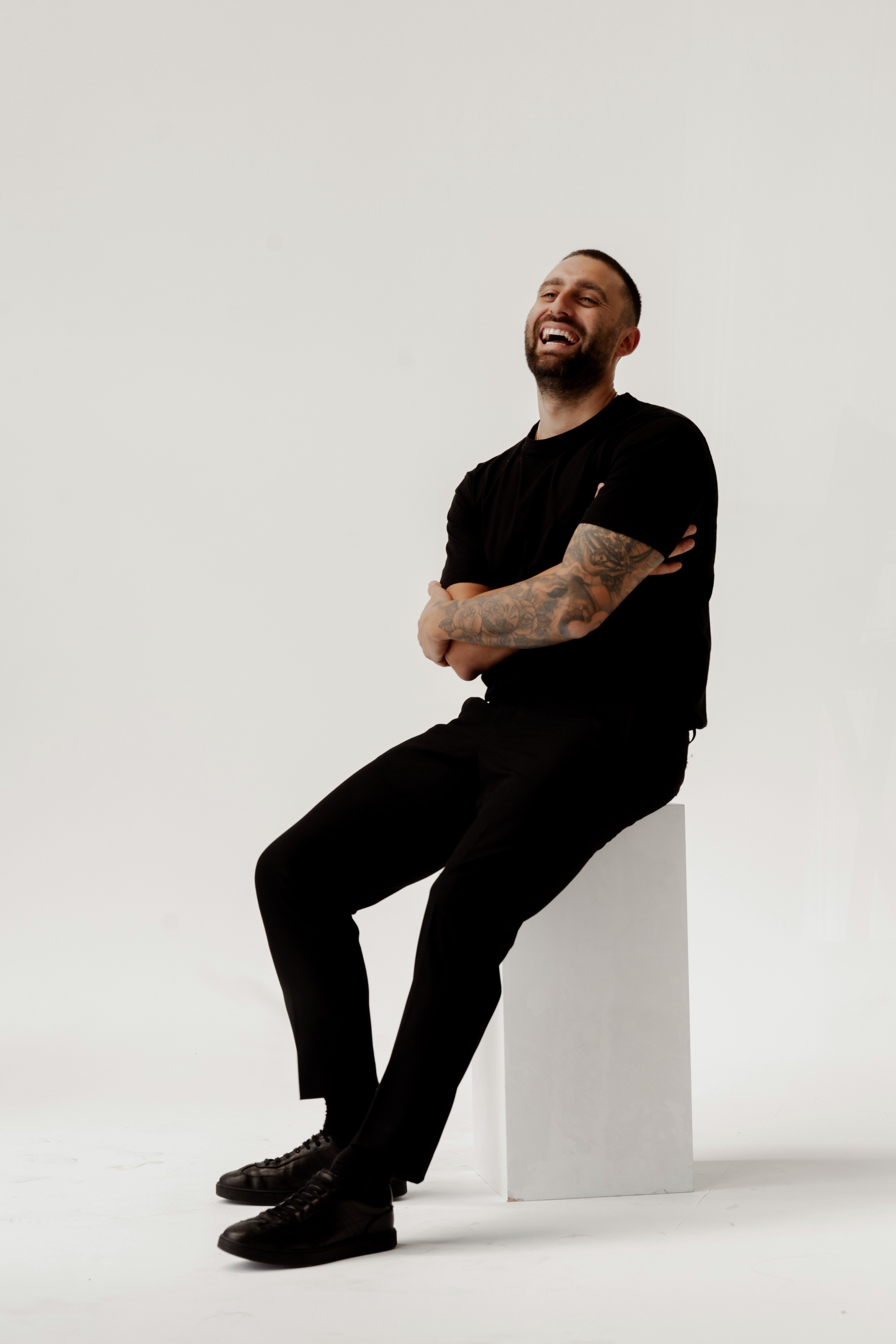 Laughing tattooed man in a black t-shirt and trousers perched on a white plinth against a bright studio backdrop.