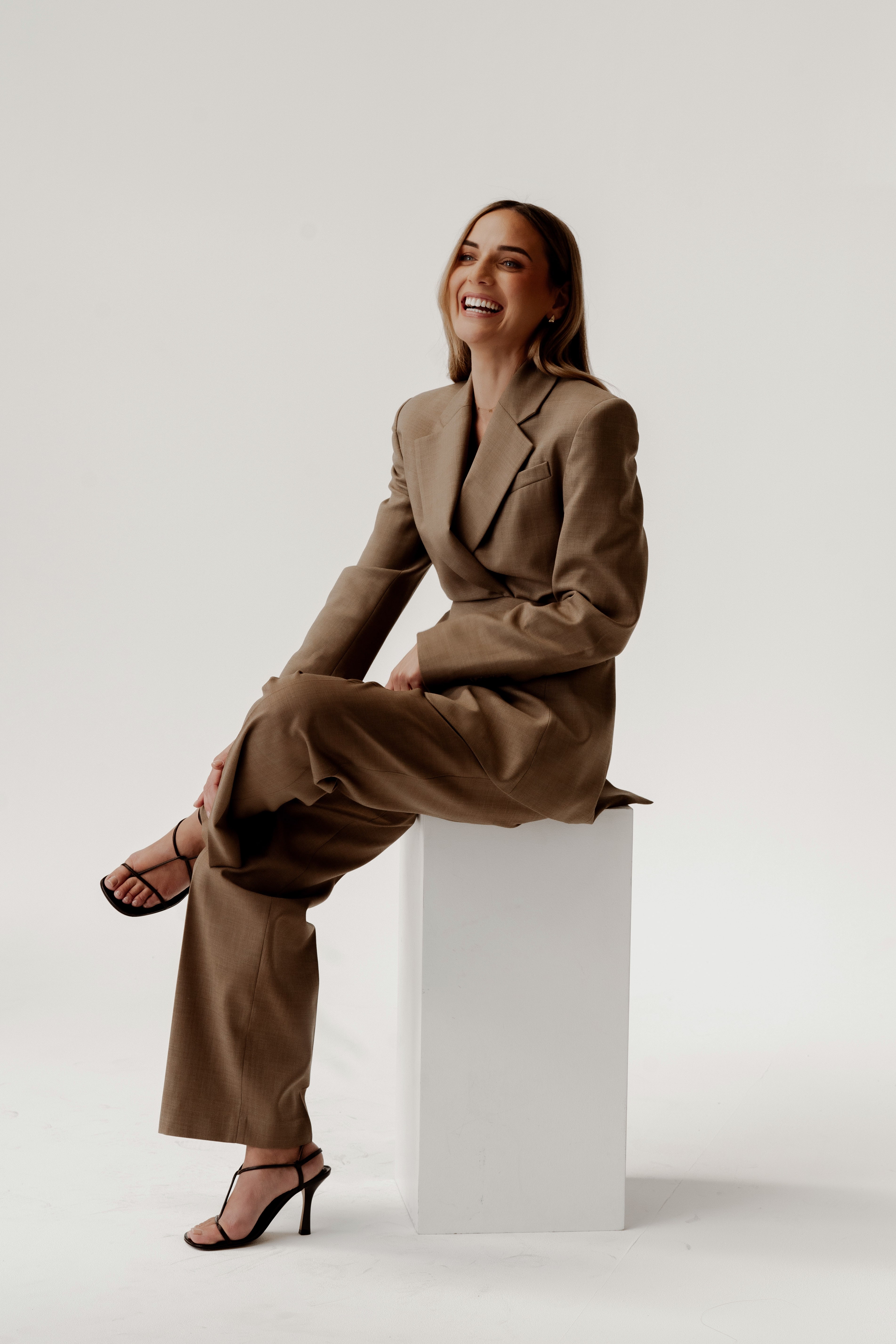 Laughing woman in a brown tailored suit and strappy heels seated on a white plinth against a light studio backdrop.