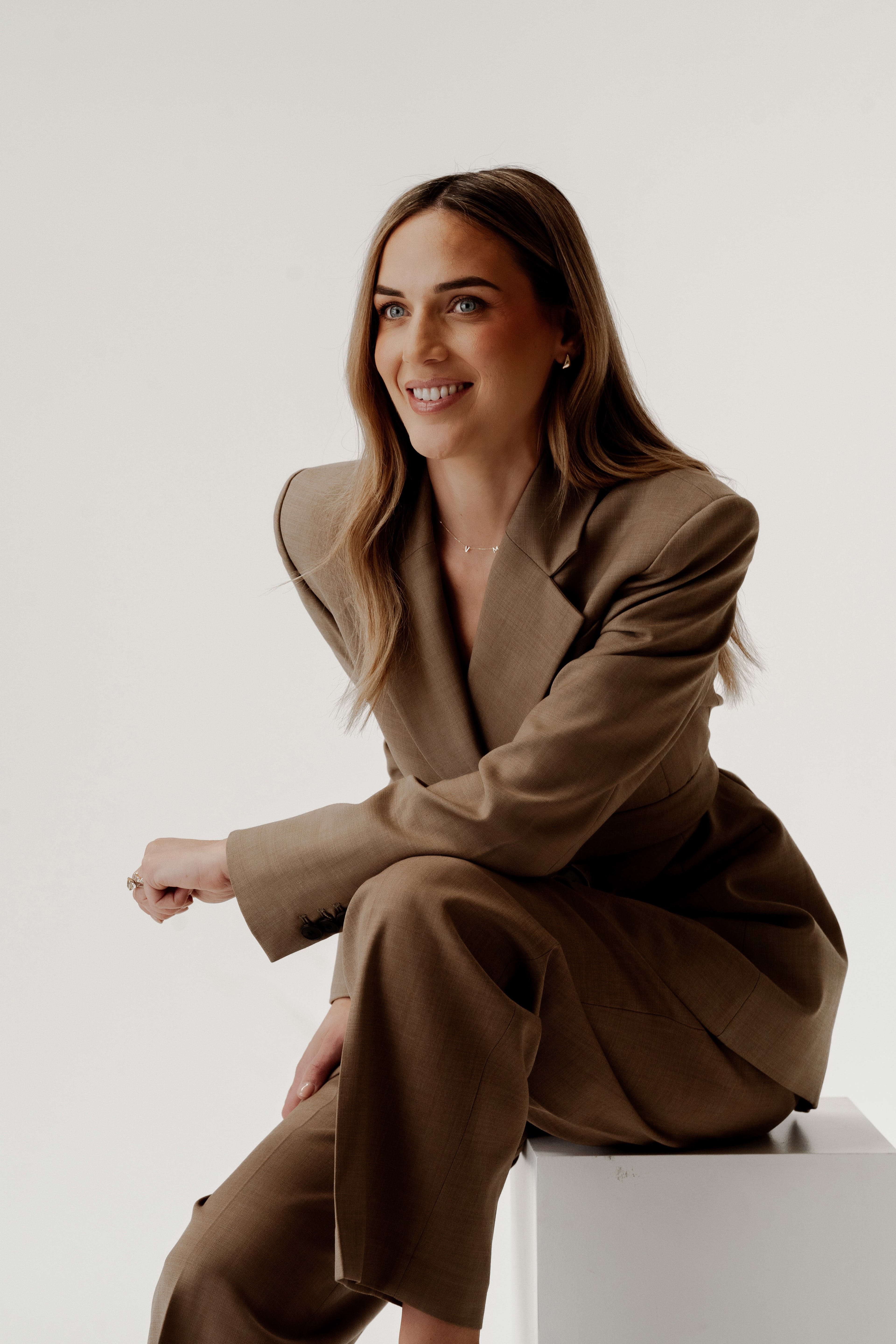 Smiling woman in a brown oversized suit seated on a white plinth against a soft neutral studio backdrop.