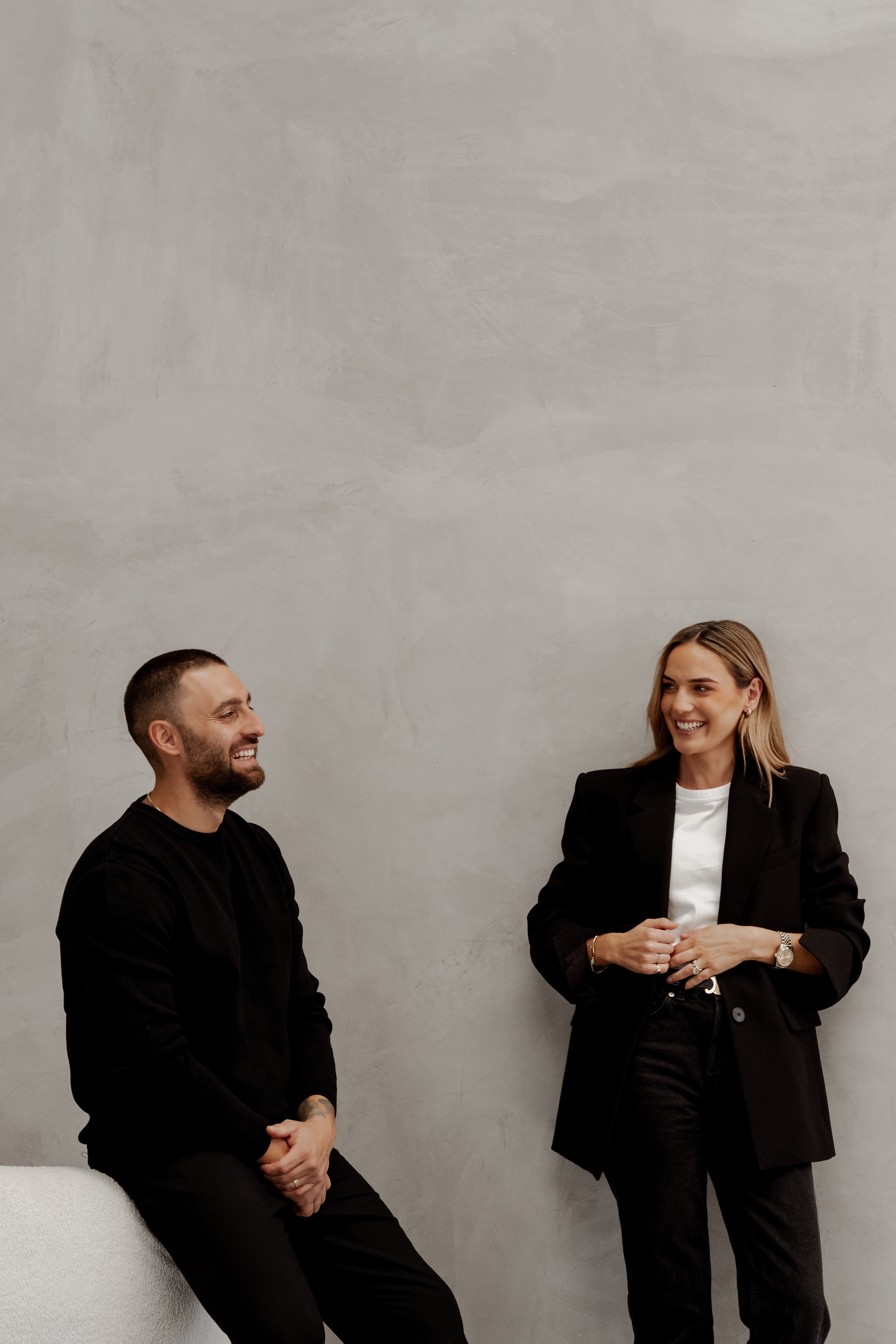 A bearded man in black and a blonde woman in a black blazer chat and smile against a textured grey plaster wall.