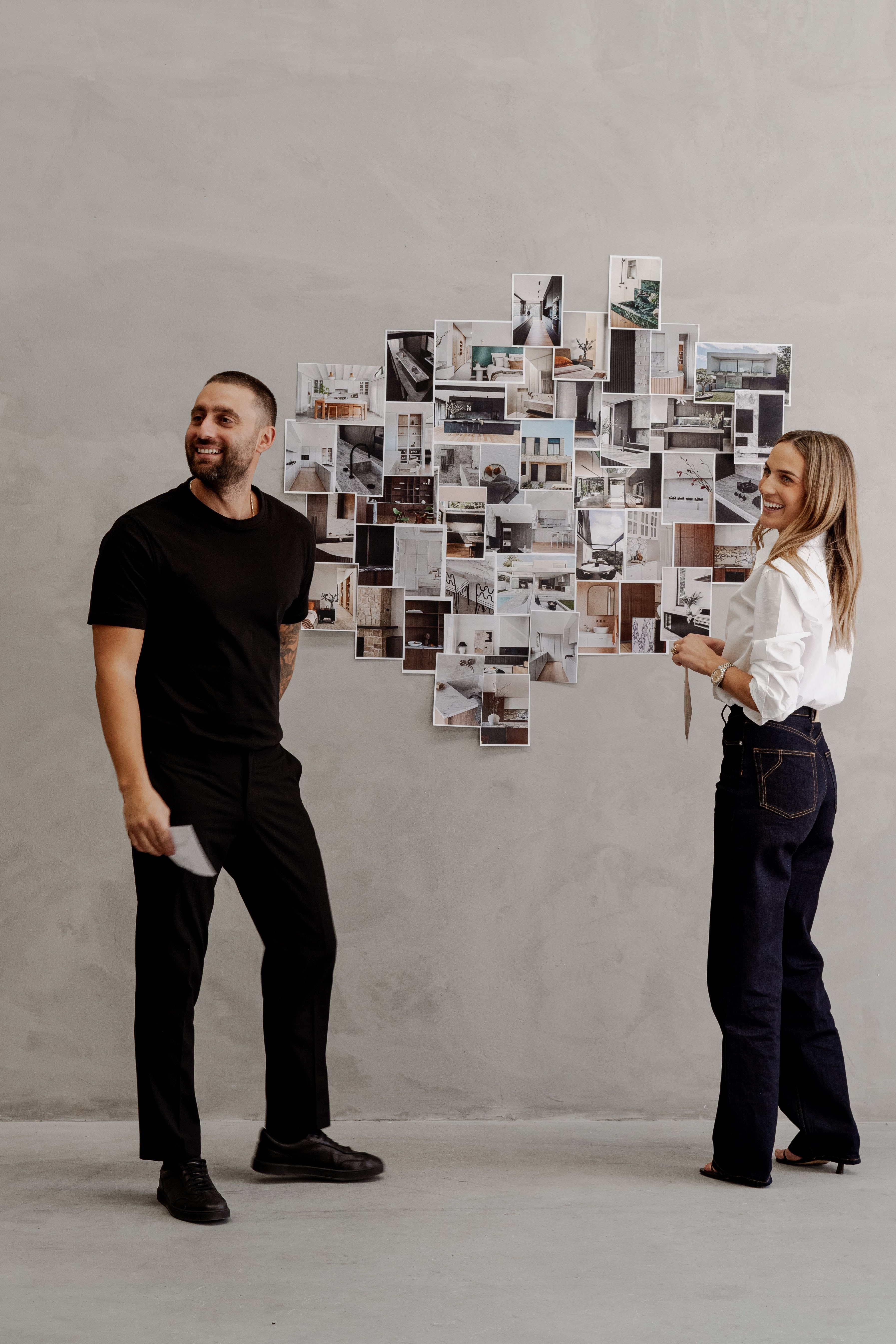 A man in black and a woman in a white shirt stand smiling either side of an interior design mood board on a grey wall.