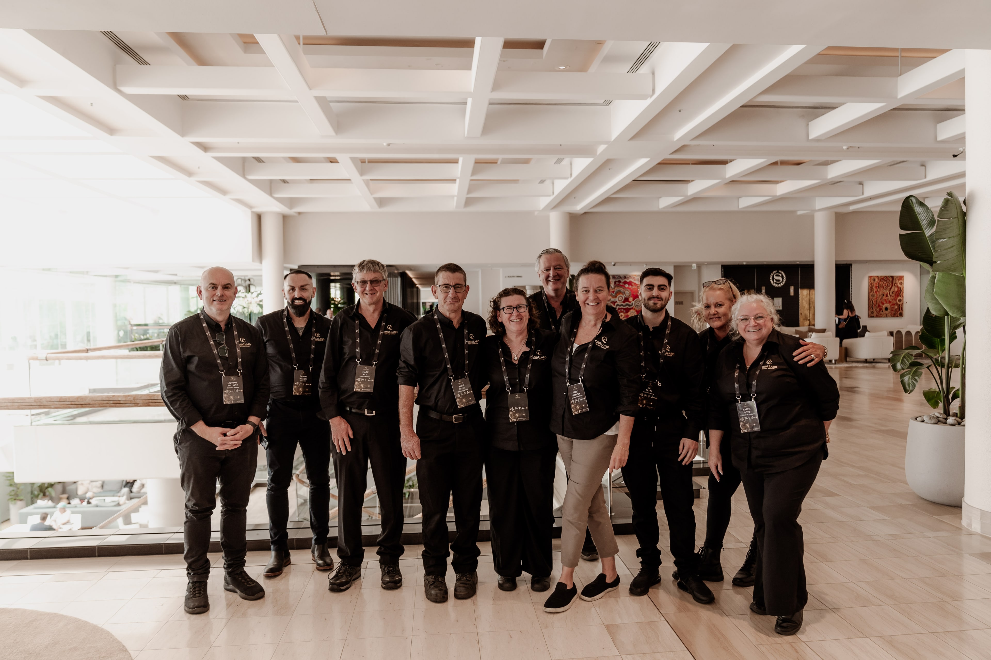 Group of eleven event staff in black uniforms and lanyards posing together in a bright resort lobby.