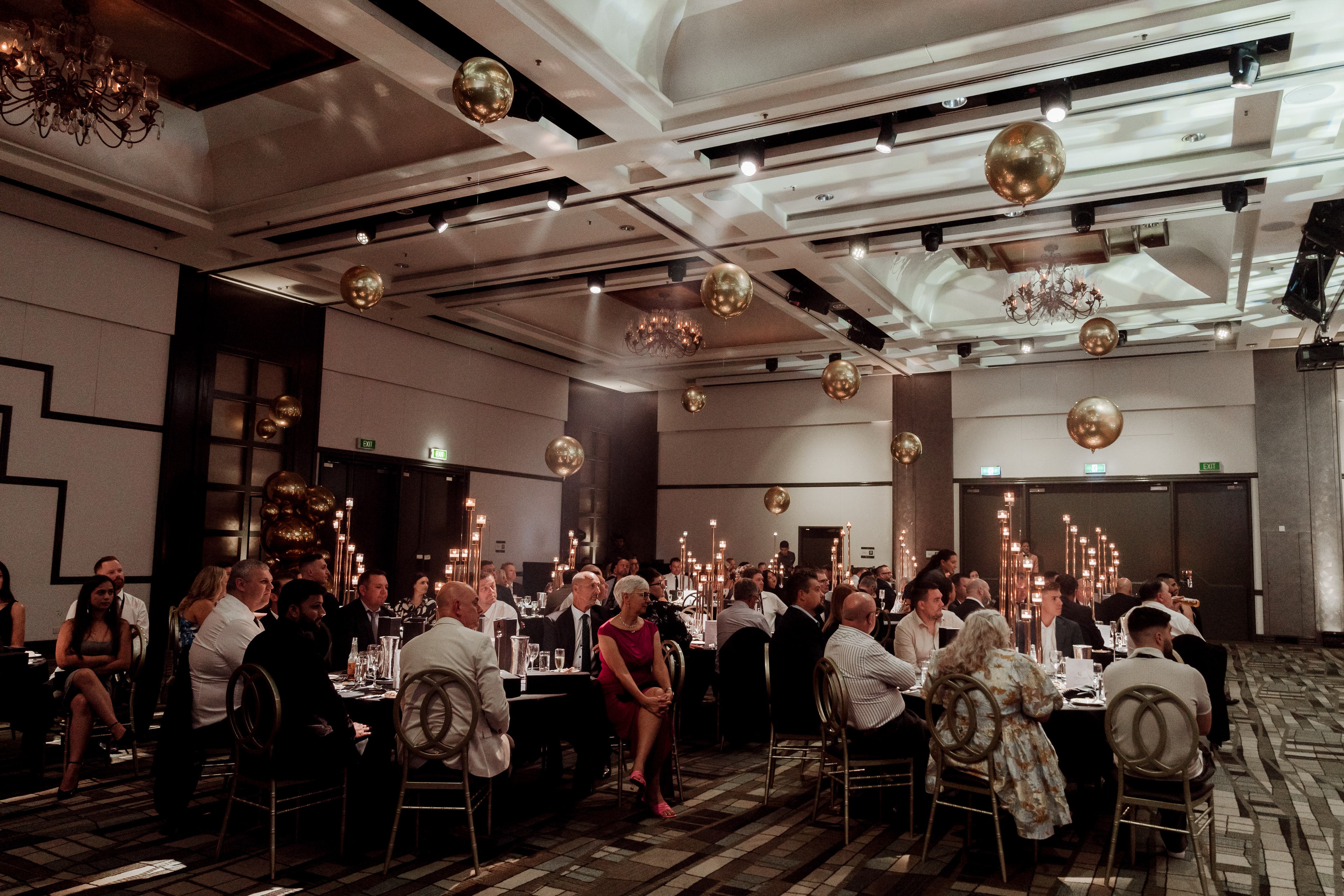 Guests seated at round banquet tables in an elegant ballroom lit by gold disco balls and tall candelabras during a gala dinner.