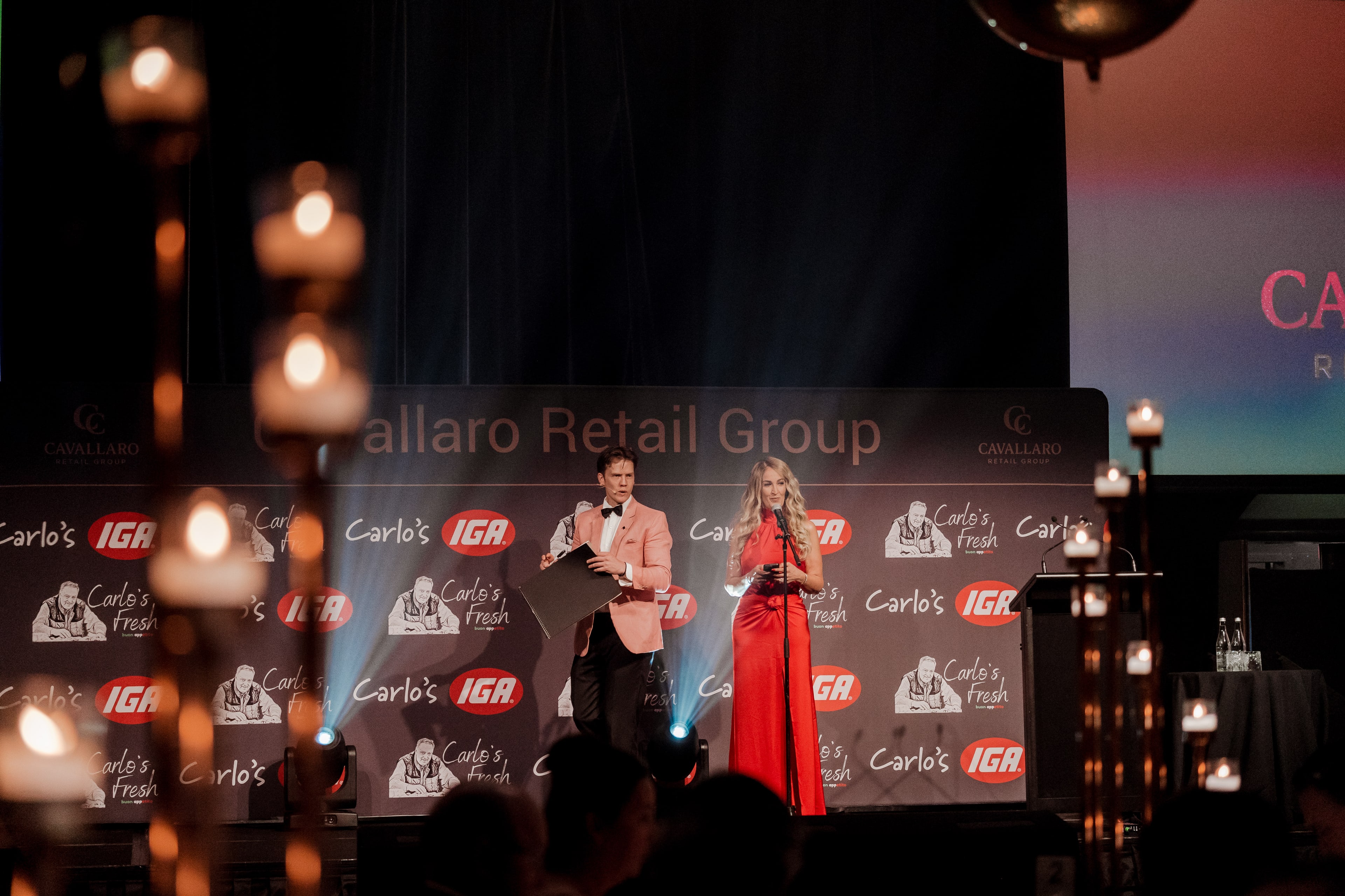 A male and female host in formal attire present on stage before a Cavallaro Retail Group and Carlo's IGA backdrop, framed by candlelight.