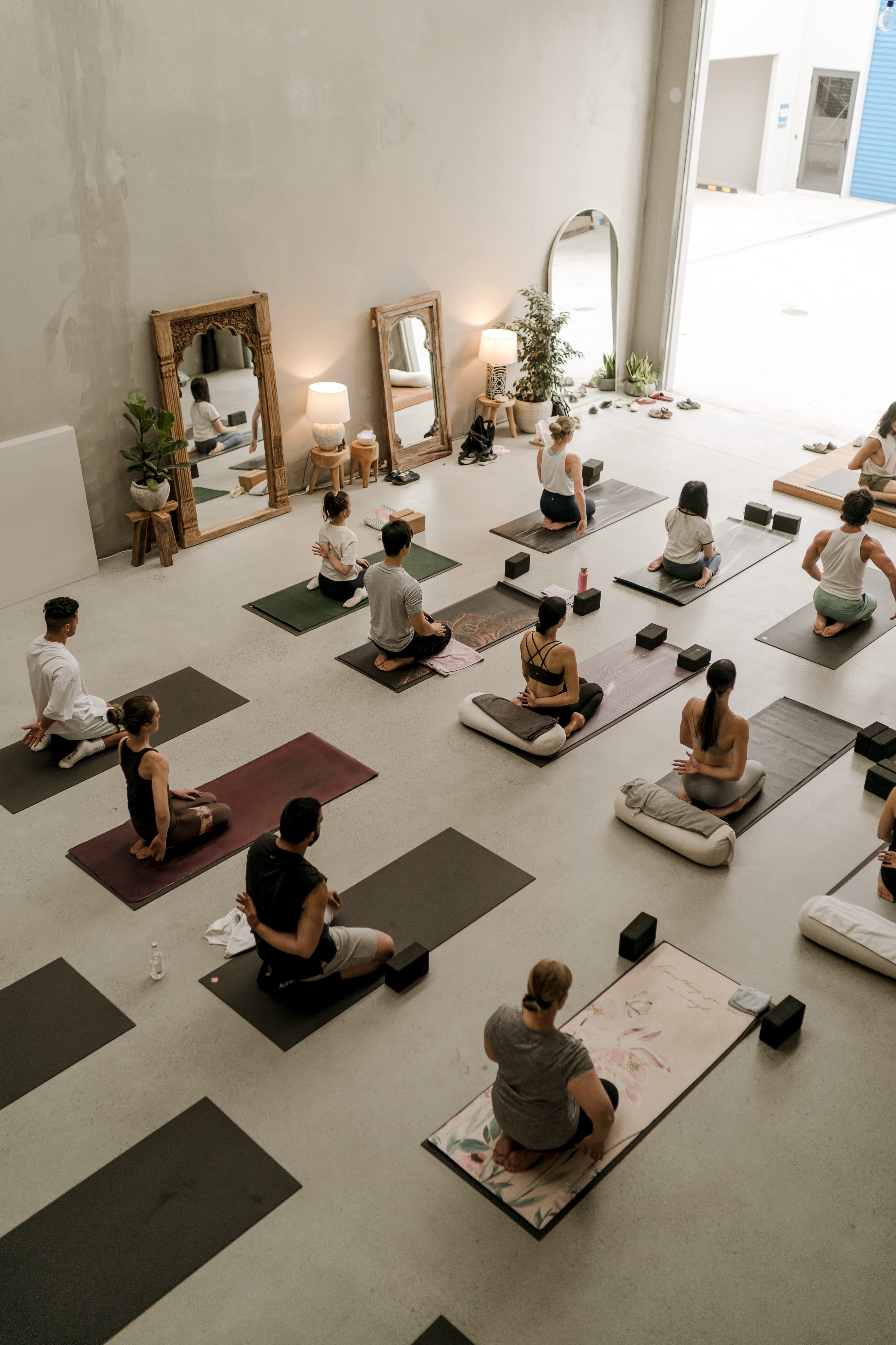 Overhead view of students seated cross-legged in meditation across a bright studio dotted with mats, cushions, blocks, mirrors and plants.