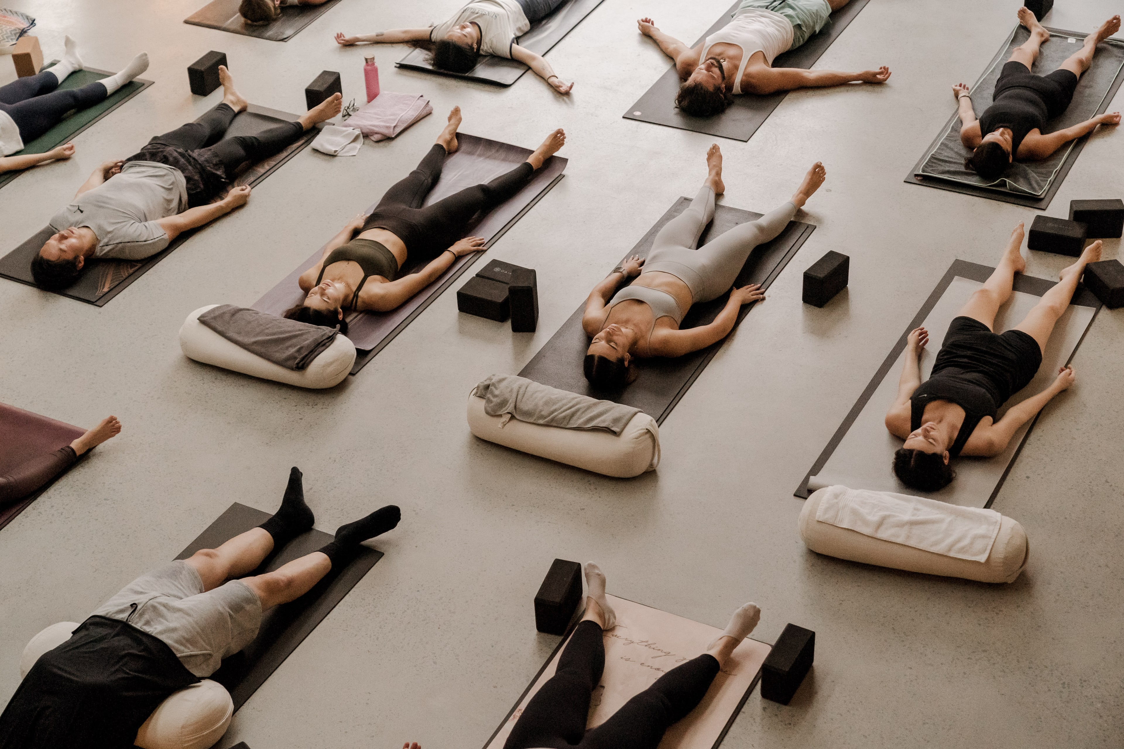Overhead view of a class lying in savasana on mats and bolsters, arms outstretched and eyes closed, on a pale studio floor.