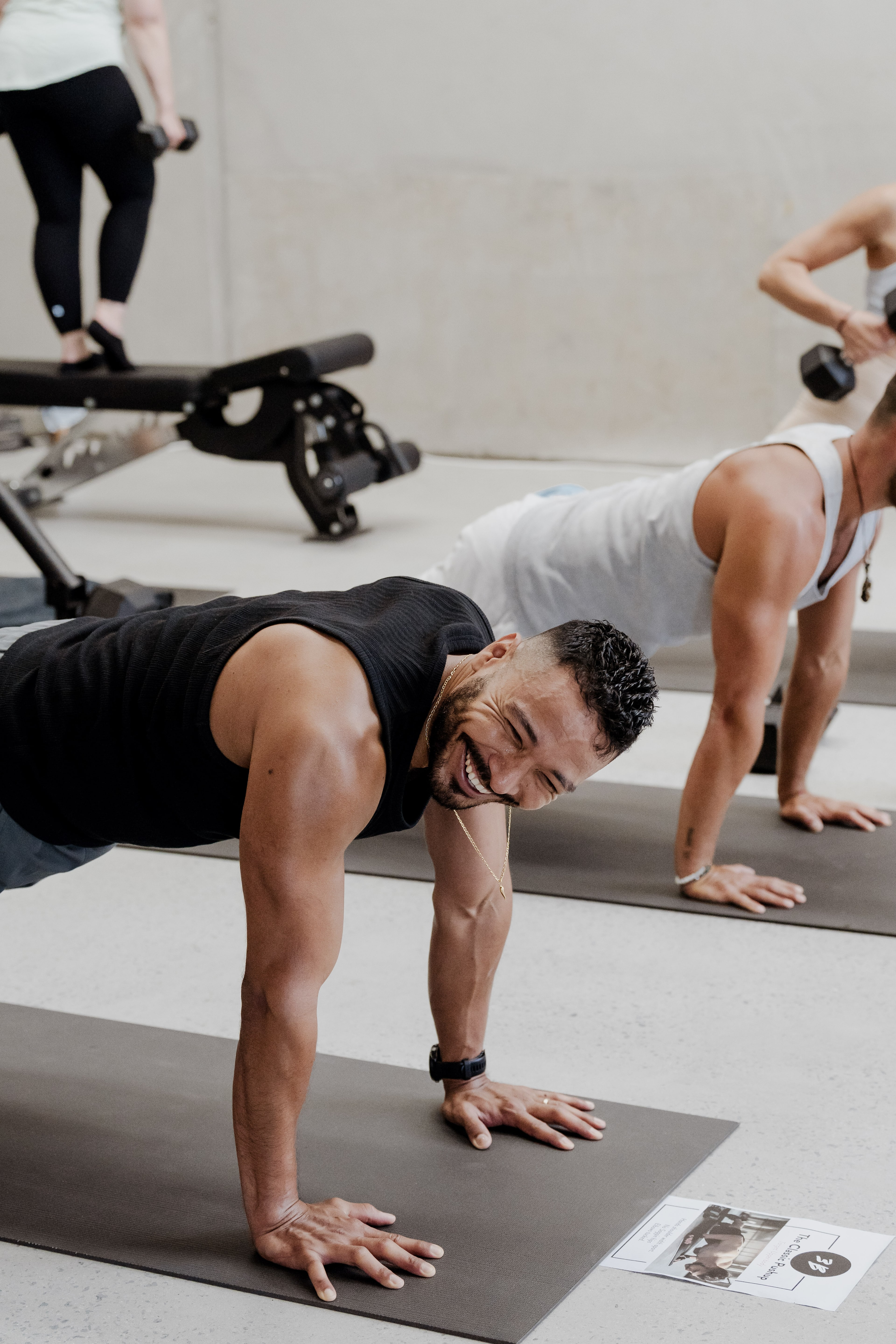 A smiling man in a black tank holds a push-up plank on a mat beside a woman lifting a dumbbell, workout benches in the background.