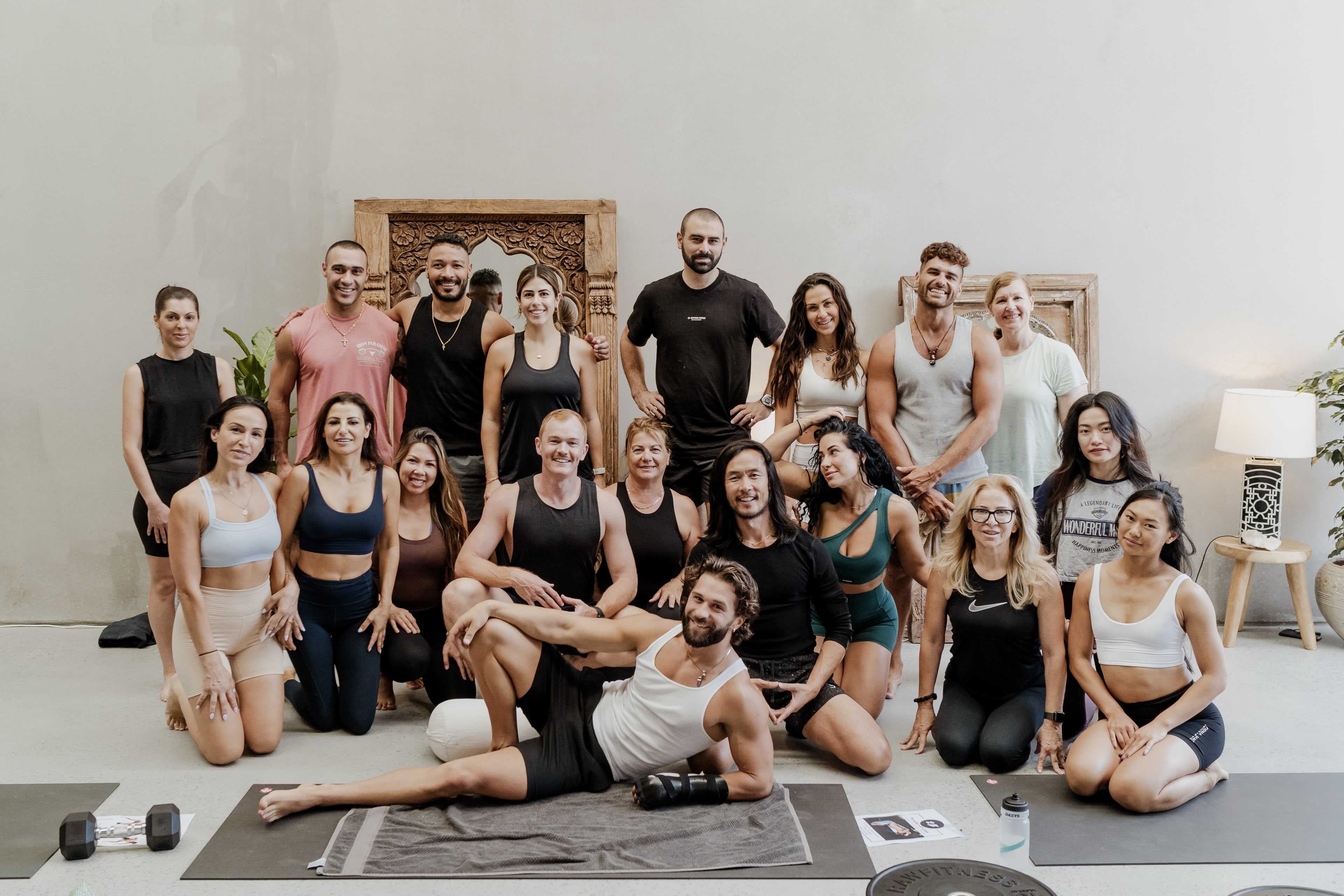 Group portrait of about twenty participants in activewear posing together in a studio before a carved wooden panel and plaster wall.