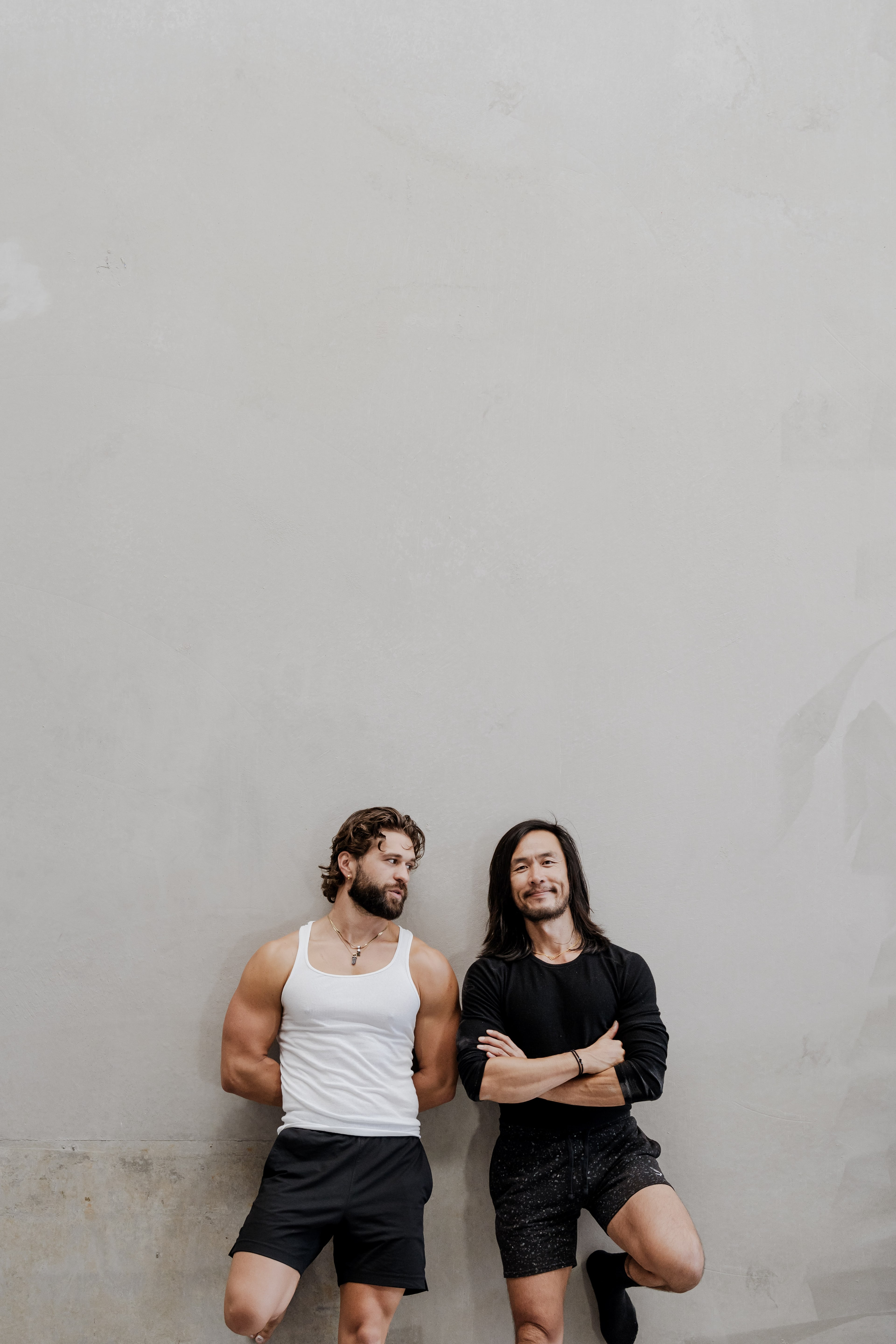Two muscular men in activewear lean against a bare concrete wall, one in a white tank and one in black with arms crossed, glancing at each other.